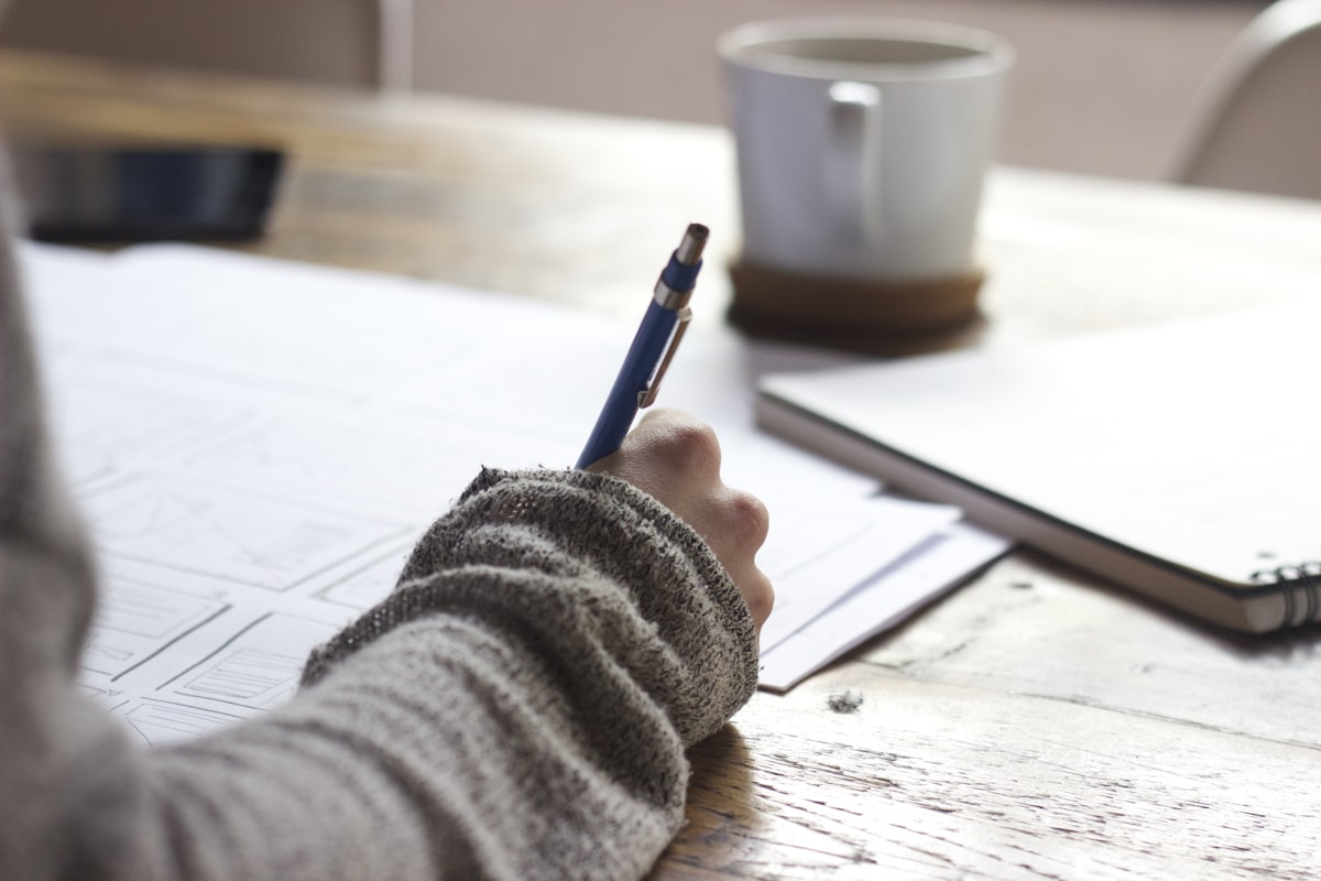 A hand holding a pen over sketched notes on paper beside a mug on a wooden desk